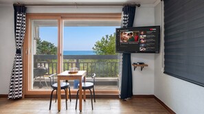 A room with a TV displaying a playlist, a table with chairs, and a view of the sea through a glass door. - Gangneung Undukmaru Pension (Gangneung)