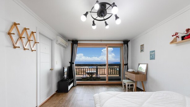 A bedroom with a bed, a desk, a chair, a TV, and a balcony with a view of the sea. - Gangneung Undukmaru Pension (Gangneung)