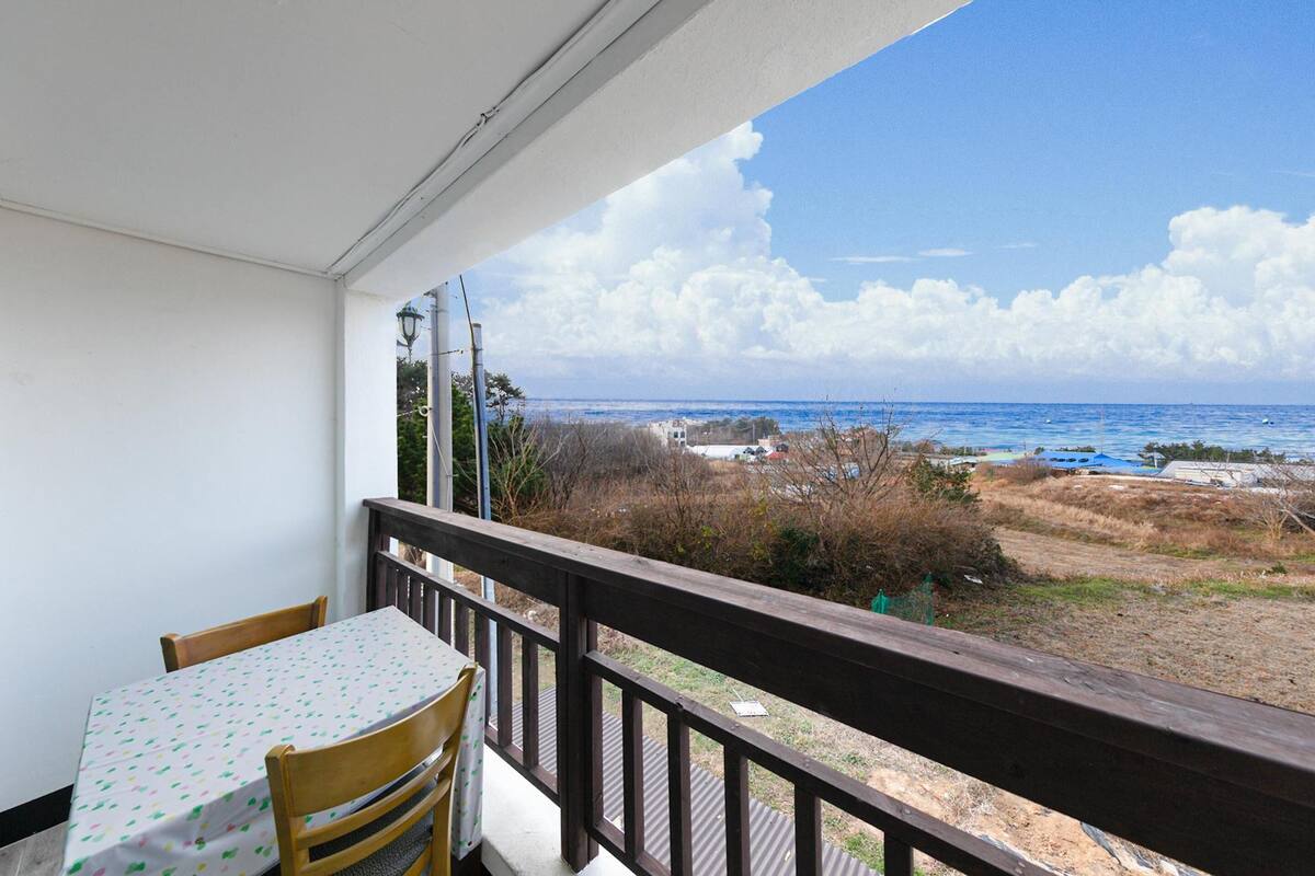 Un balcon avec une table et des chaises, donnant sur une plage, avec un ciel dégagé et des bâtiments visibles au loin.