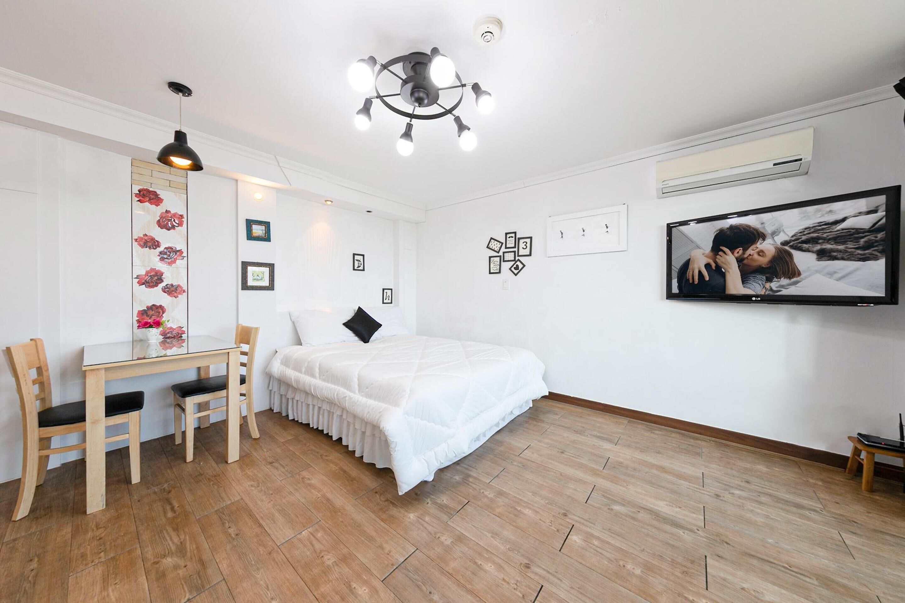 A bedroom with a bed, a desk with chairs, a wall-mounted TV, and a ceiling light fixture.