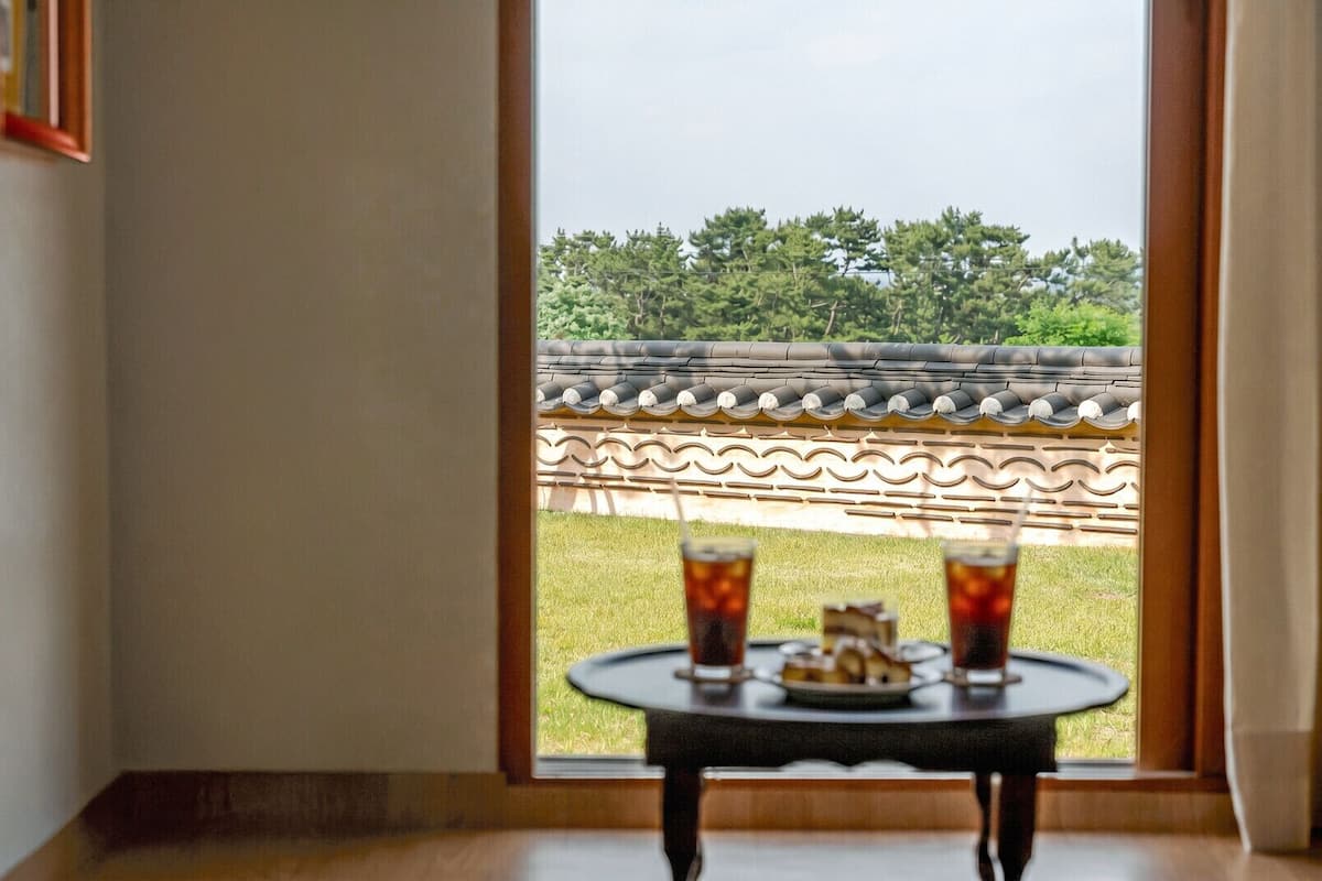 A room with a view of traditional Korean architecture, a small table with drinks, and a framed picture on the wall.