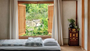 A bedroom with a bed, a window with wooden shutters, a dresser with decorative elements, and a view of greenery outside.