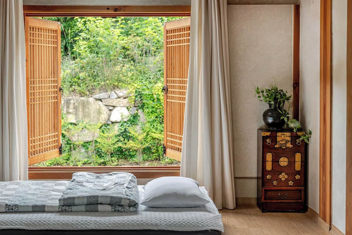 A bedroom with a bed, a window with wooden shutters, a dresser with decorative elements, and a view of greenery outside.