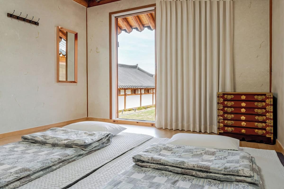 A room with two beds, a wooden floor, a window with a view of a traditional building, and a wooden cabinet with drawers.