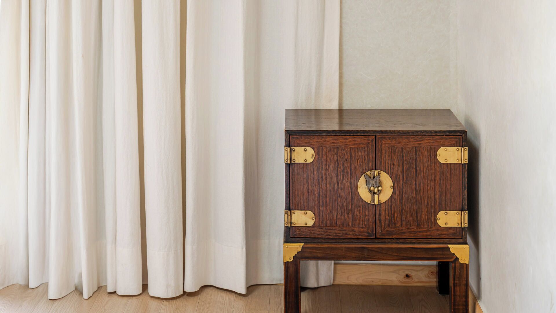 A wooden cabinet with brass hinges and a circular handle, placed against a wall with a curtain.
