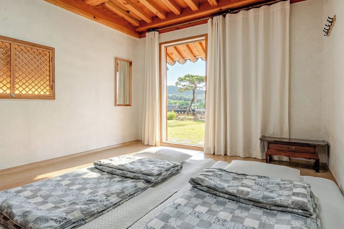 A bedroom with two beds, a wooden ceiling, a window with curtains, and a view of the outdoors.