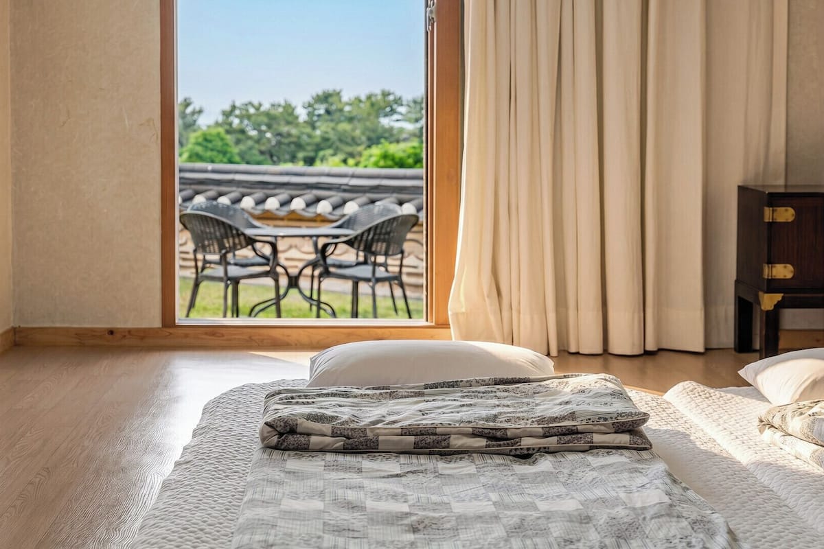 A bedroom with a large window, a bed with a patterned bedspread, a wooden floor, and a view of an outdoor dining area.