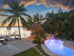 A pool area with a thatched umbrella, palm trees, and a view of the water. - Key West Beach House with heated pool, tiki, and dock.♿︎ (Key West)