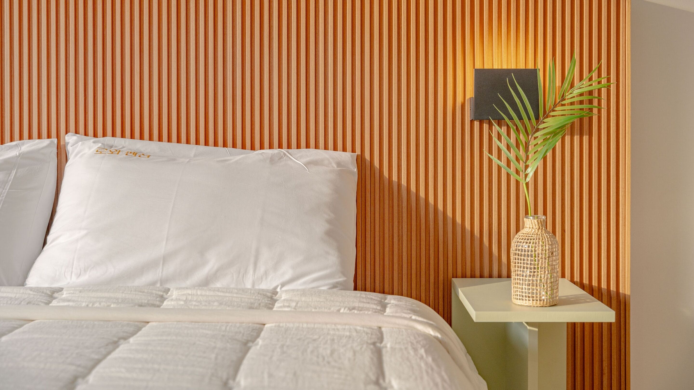 A neatly made bed with white bedding, a small bedside table with a woven vase, and a vertical wooden panel wall.