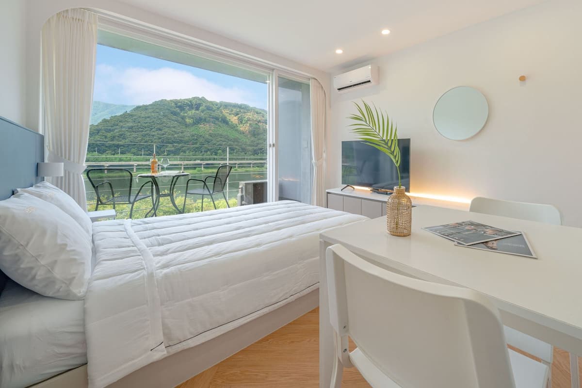 A hotel room with a bed, a desk, a TV, and a view of a green landscape.