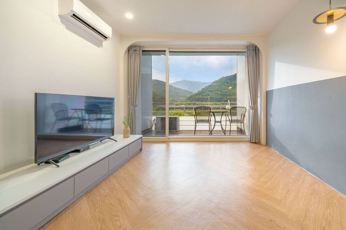 A modern living room with a flat-screen TV, a small table with chairs, and a balcony with a scenic mountain view.
