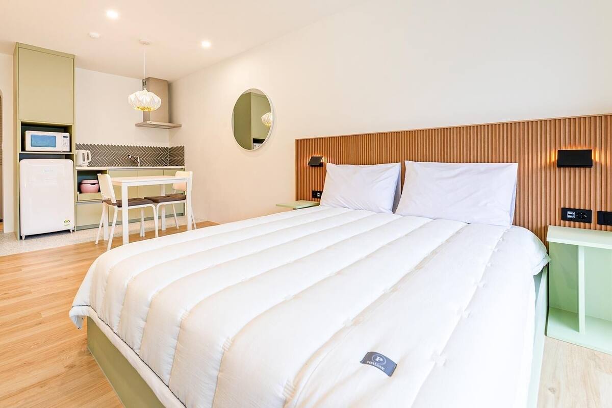 A modern hotel room with a bed, bedside table, and a small kitchenette.