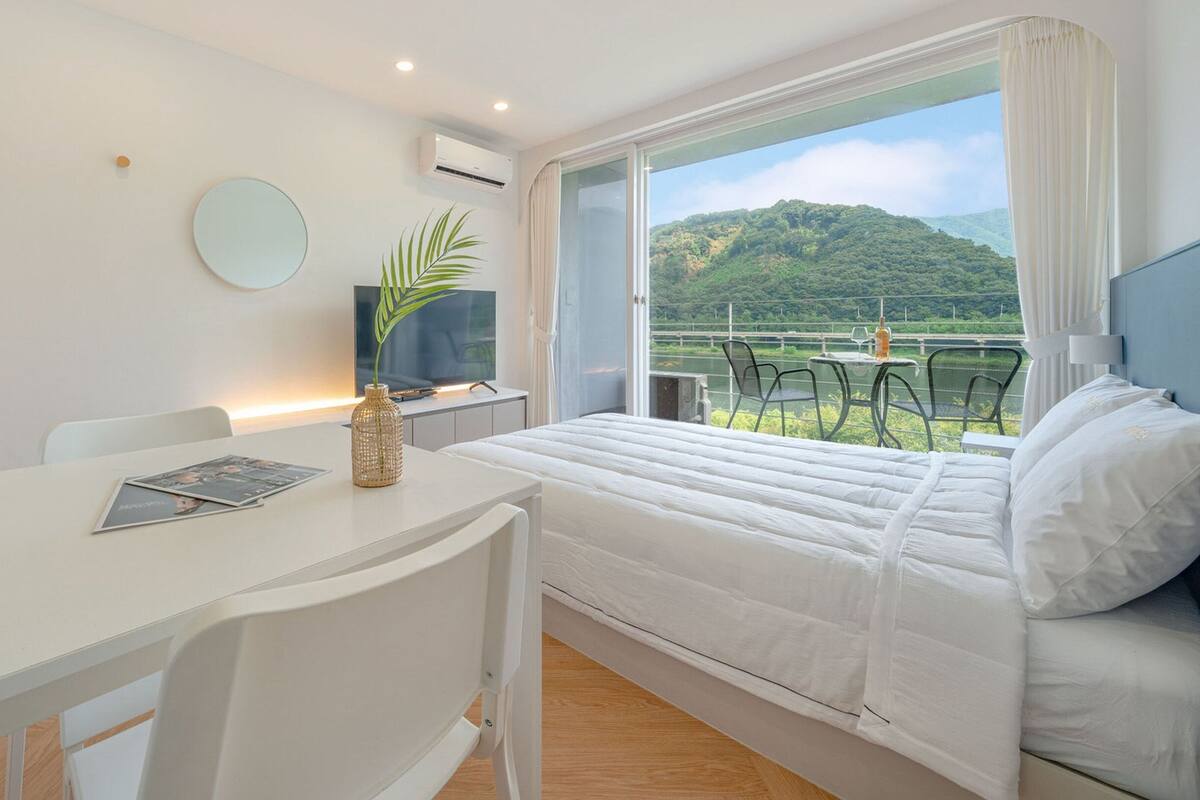 A hotel room with a bed, a desk, a TV, and a view of a green landscape through the window.