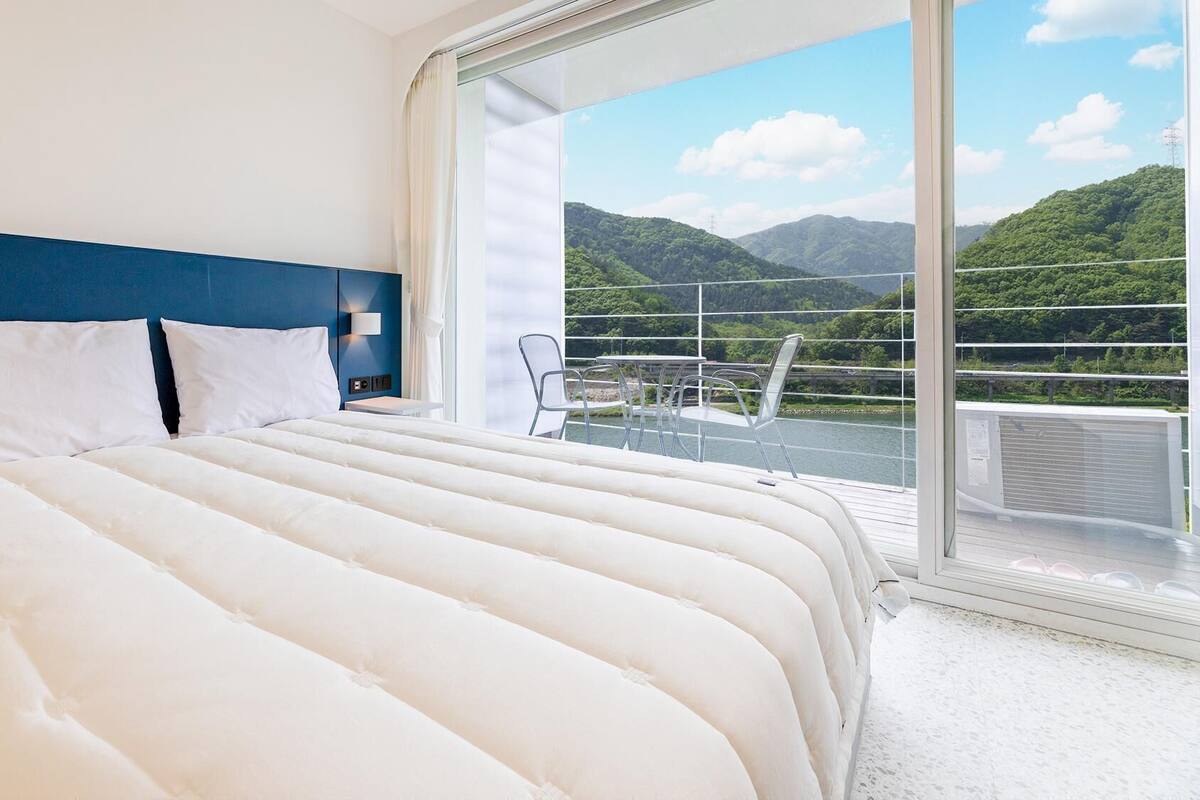 A hotel room with a large bed, a nightstand, a lamp, and a view of a balcony with a railing and mountains in the background.