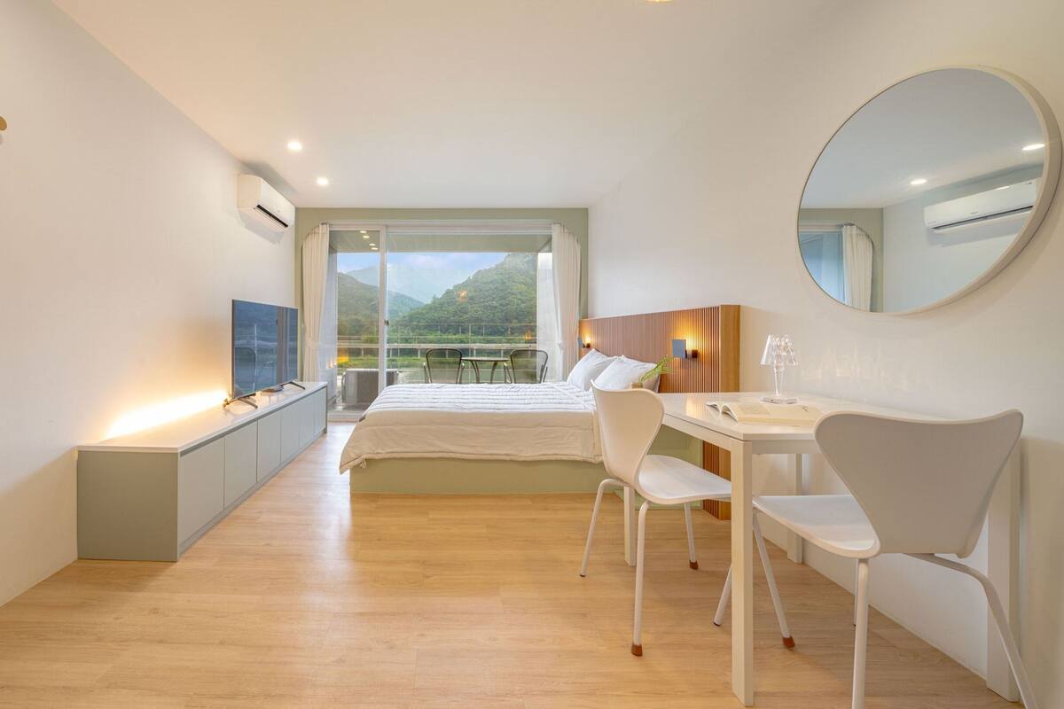 A modern hotel room with a bed, a desk, a chair, a TV, and a large window offering a mountain view.