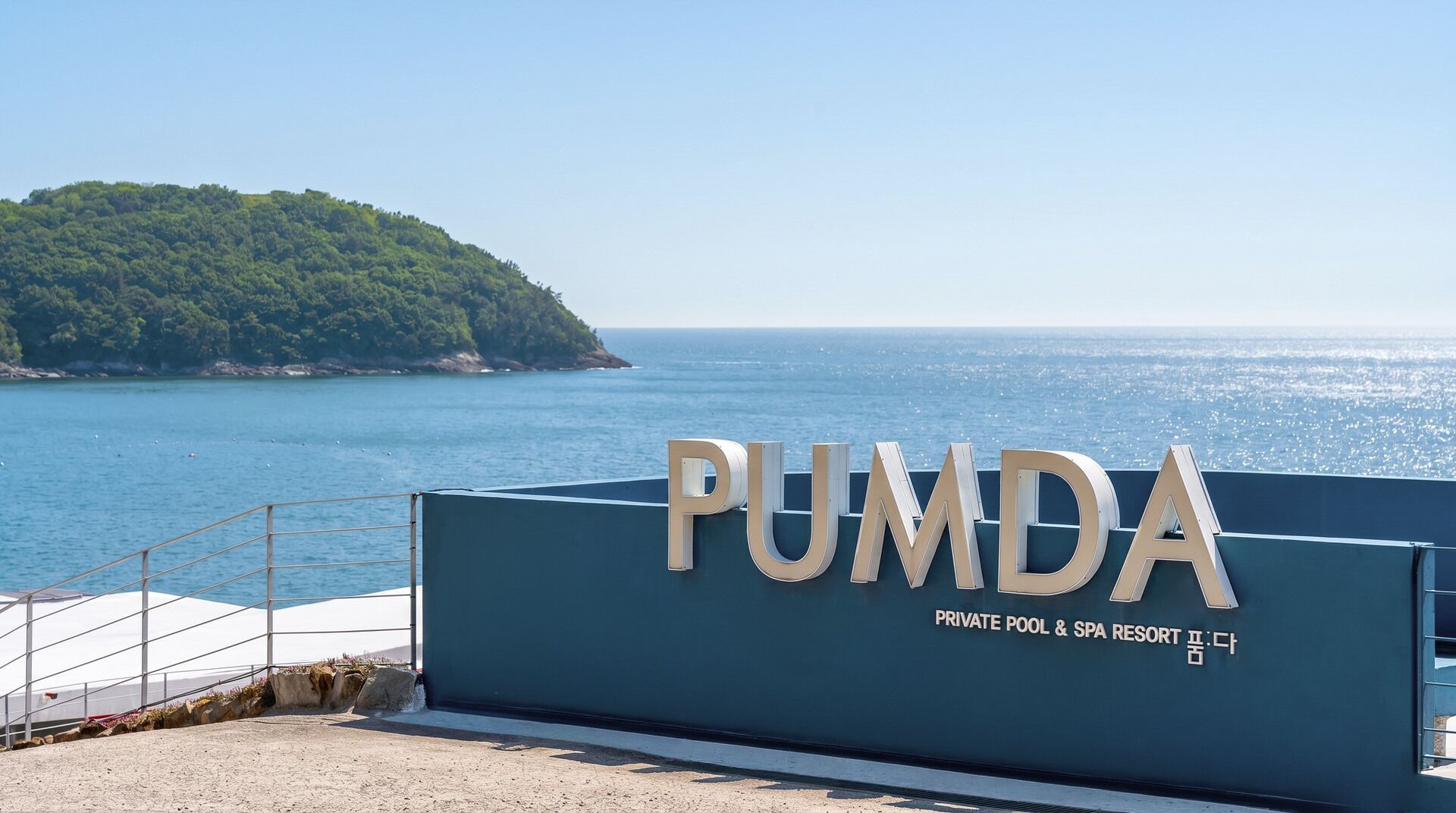 Letrero que dice 'PUMDA PRIVATE POOL & SPA RESORT', con vista al mar y una colina verde al fondo.