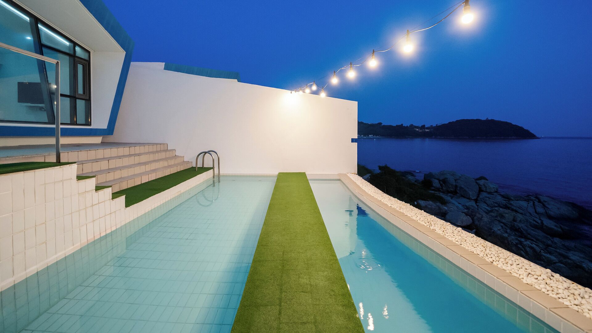 Una piscina con alfombra verde, escalones que llevan a una puerta de cristal y luces colgantes contra un cielo crepuscular.