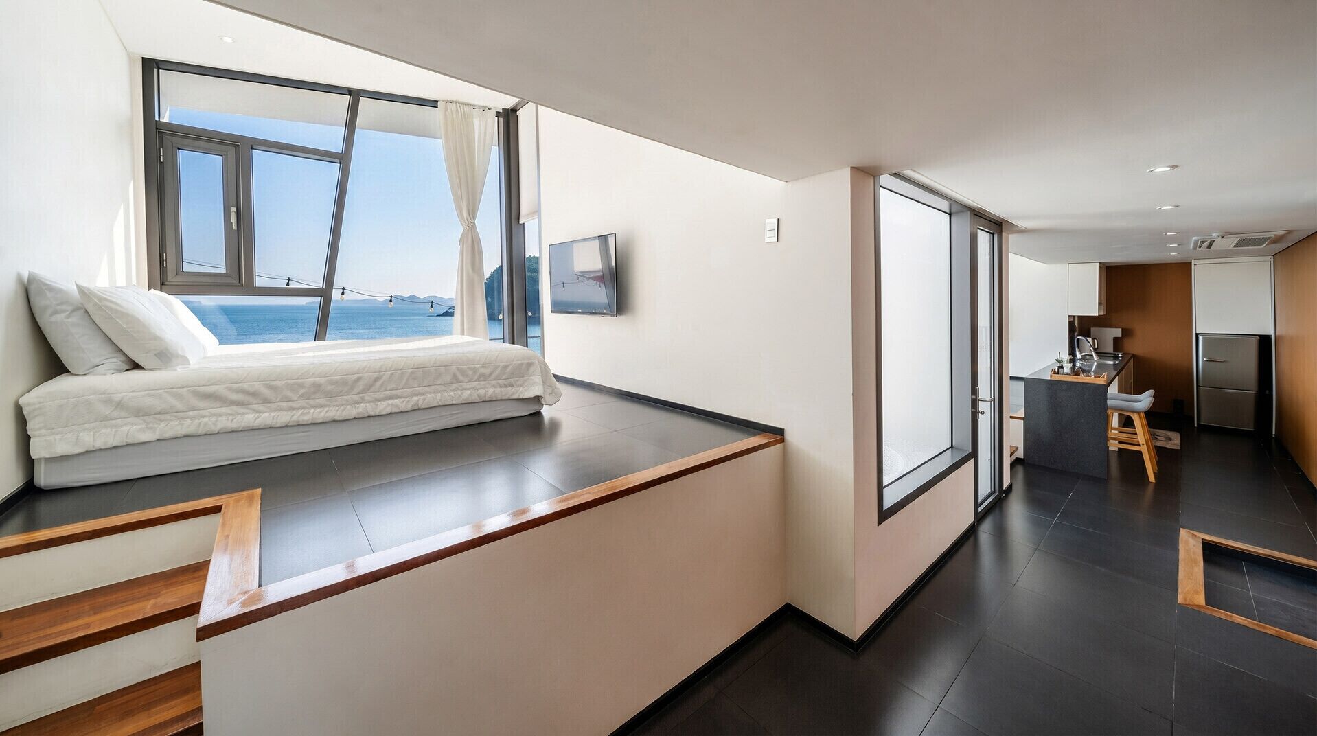 Une chambre moderne avec un lit, une grande fenêtre avec des rideaux, une vue sur la mer et un coin cuisine équipé d’un réfrigérateur et d’un plan de travail.