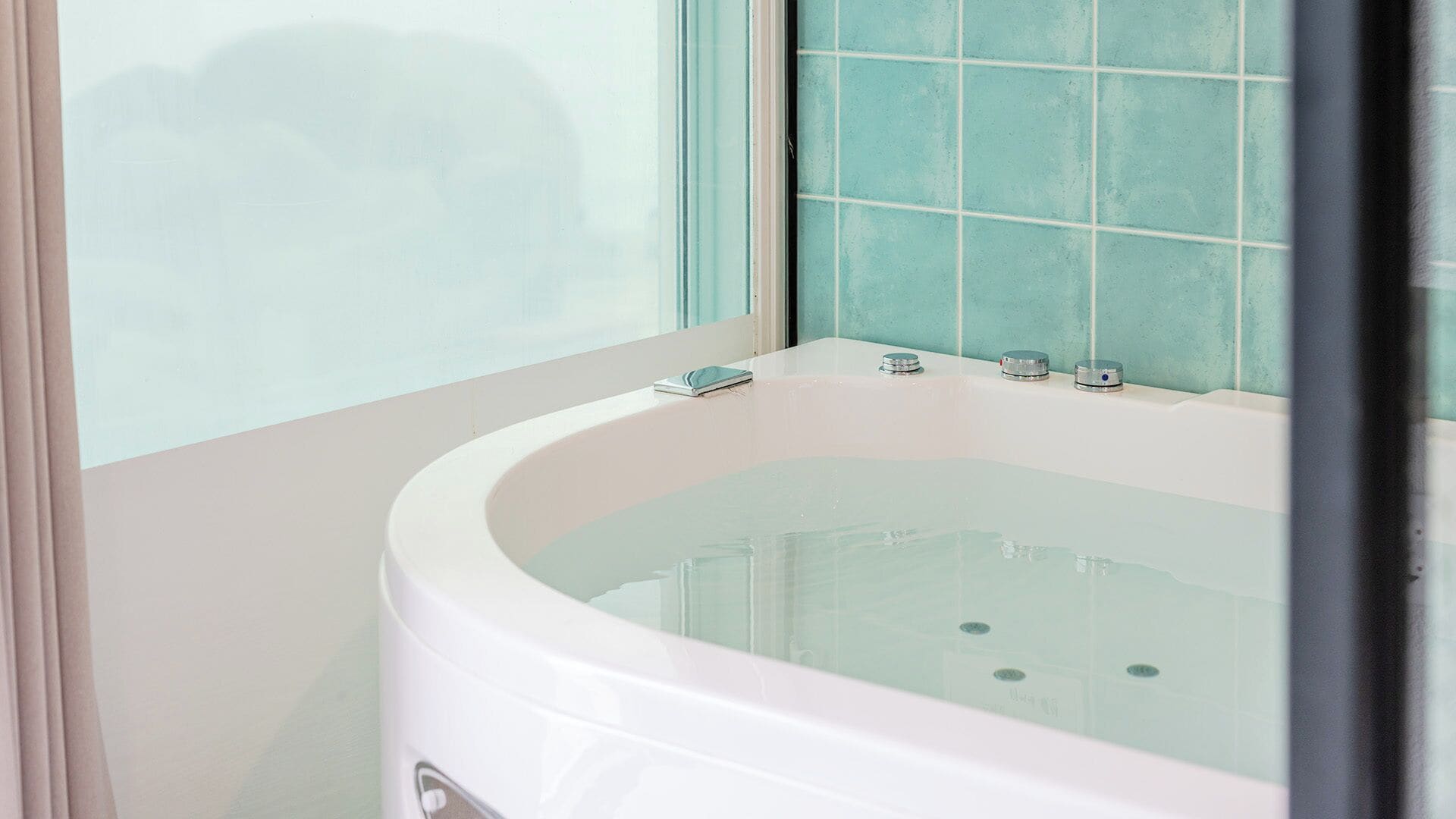 Eine Badewanne mit Glasabtrennung, ein Fenster mit satinierten Scheiben und eine geflieste Wand.