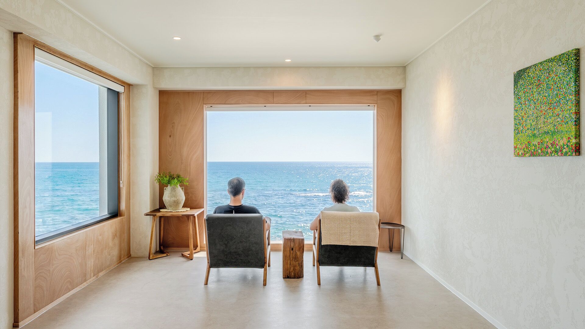 A modern living room with a large window overlooking the sea, a small wooden table with a plant, and a minimalist design.