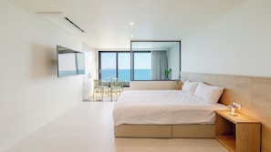 Une chambre d’hôtel moderne avec un grand lit, une table de chevet avec une lampe, un téléviseur fixé au mur et une vue sur la mer grâce à de grandes fenêtres.