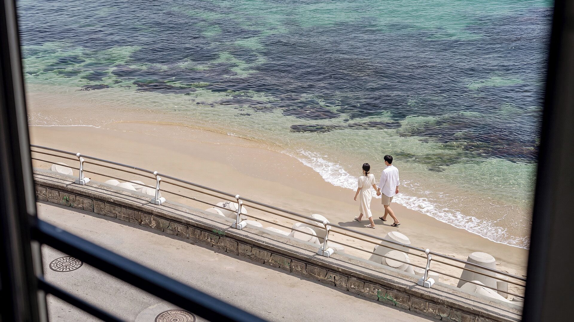 A couple walking on a beach with a railing and a view of the ocean.