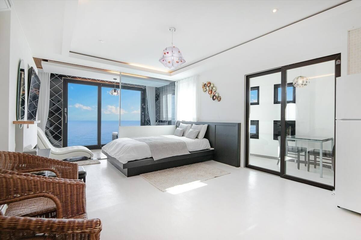 Un dormitorio moderno con una cama, un ventanal de vidrio que da a un balcón y un refrigerador.