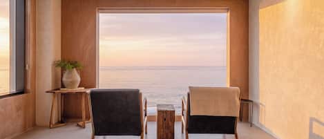 Une pièce dotée d’une grande fenêtre donnant sur la mer, de deux fauteuils, d’une petite table et d’une plante.