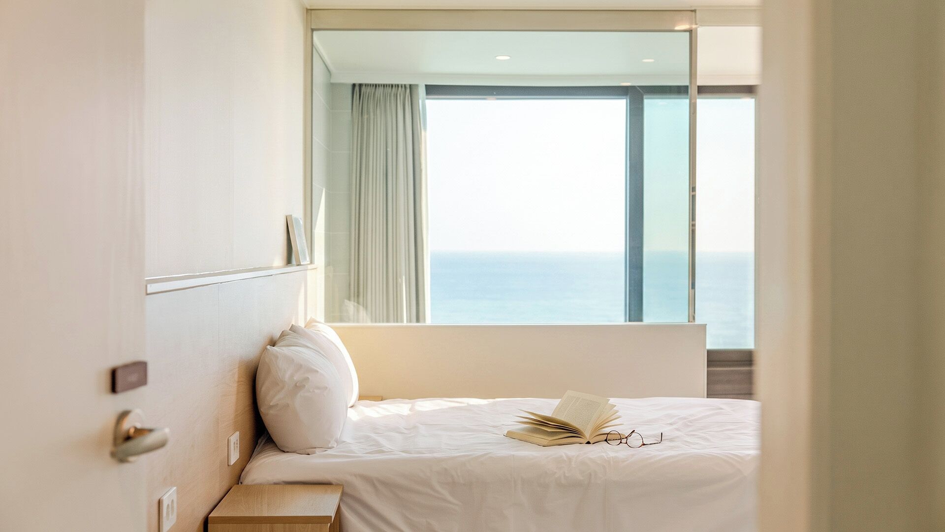 A hotel room with a bed, a bedside table, a nightstand, a large window with a view of the sea, and a mirror on the wall.