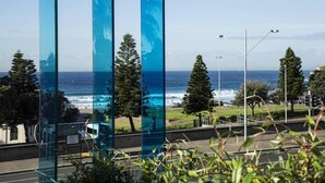 Une vue sur la côte, avec des panneaux de verre qui reflètent l’océan, les lampadaires et les arbres.