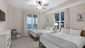 Habitación de hotel con dos camas, un escritorio, una silla, un ventilador de techo y ventanas grandes con cortinas.