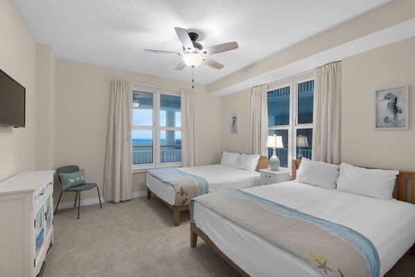 A hotel room with two beds, a desk, a chair, a ceiling fan, and large windows with curtains.