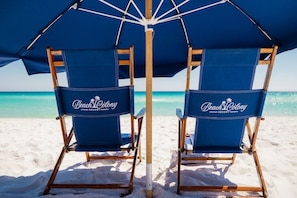 Deux chaises de plage bleues, portant le logo « Beach Colony Resort », sont placées sous un grand parasol bleu, sur une plage de sable offrant une vue dégagée sur l’océan.