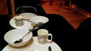 En matplats med ett bord dukat för två personer, inklusive tallrikar, skålar, en mugg och bestick. Det finns två stolar, en grön och en röd, och ett litet bord med en stol i bakgrunden.