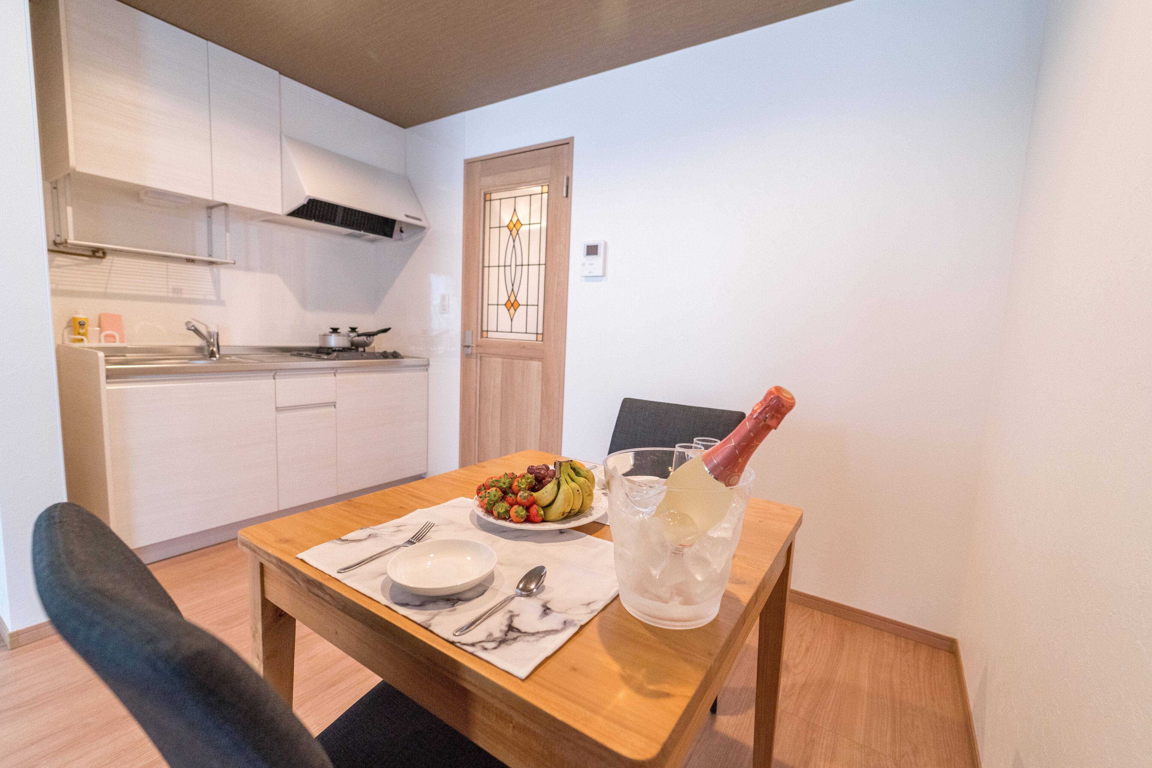 Una cocina compacta con armarios blancos, una mesa de comedor de madera con una botella de champán y un bol de fruta.