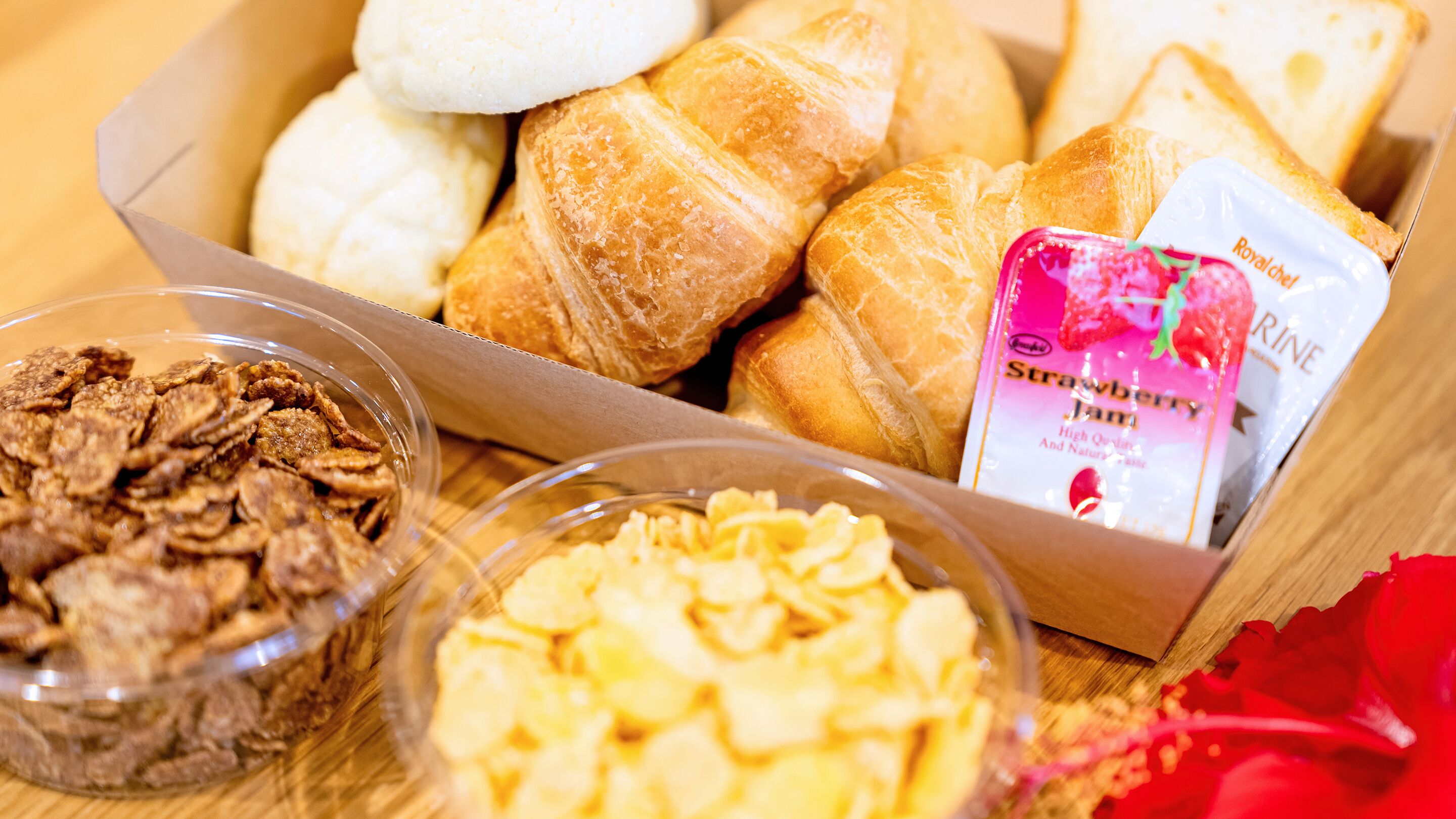 A box containing croissants, a packet of strawberry jam, and two bowls with cereal and puffed rice.