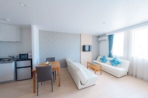 A modern living room with a sofa, a dining table, and a kitchen area with a microwave and refrigerator. - Hotel California Miyakojima Resort (Miyakojima)