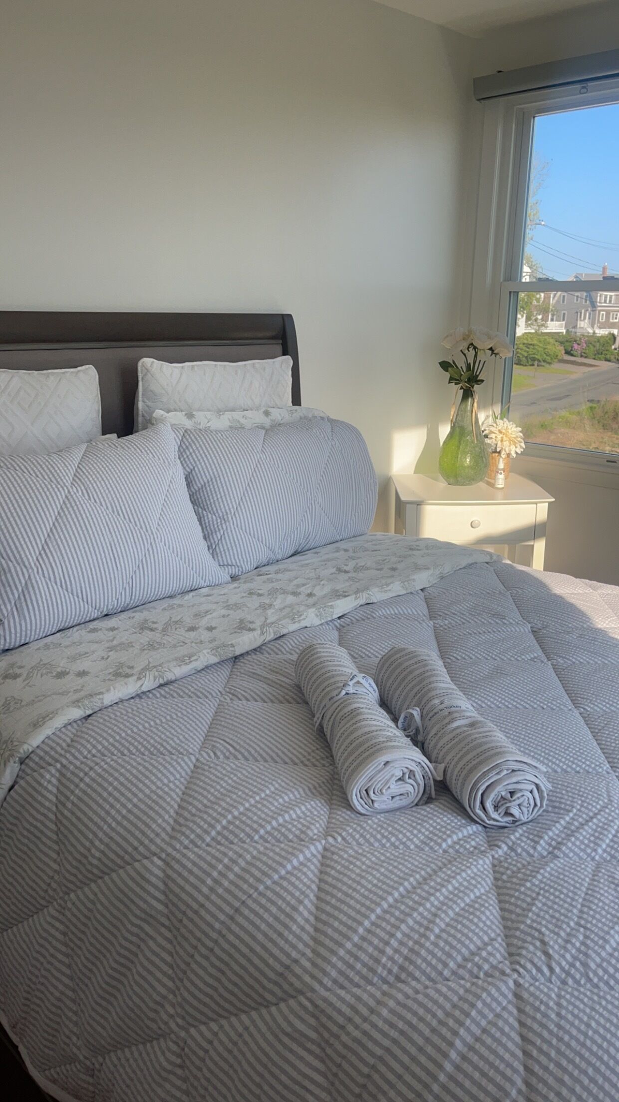 A neatly made bed with striped bedding and a rolled-up blanket, a vase with flowers on a bedside table, and a window with a view of houses.