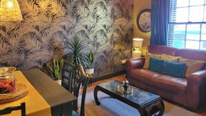 Ein Wohnzimmer mit strukturiertem Wandpapier, einem Ledersofa mit Kissen, einem Glastisch, einem Esstisch mit Stühlen, einer Topfpflanze und einem Fenster mit Vorhängen.