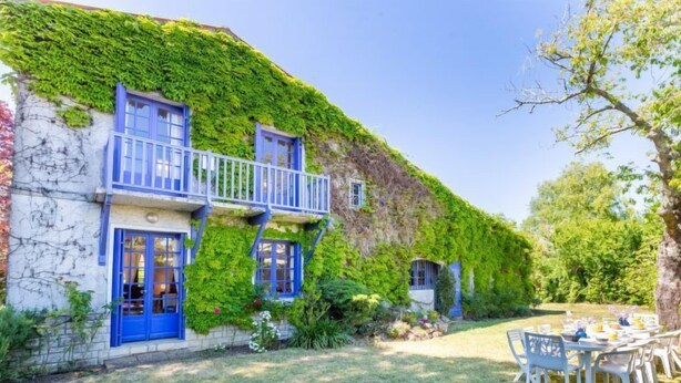 Un bâtiment à la façade imposante, recouverte de lierre, doté d’un balcon bleu et d’une cour verdoyante aménagée avec des sièges extérieurs.