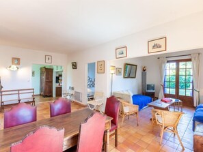 Un salon spacieux avec une table à manger en bois, des chaises rouges et blanches, un canapé bleu et un téléviseur. Les murs sont ornés de tableaux encadrés et une grande fenêtre est agrémentée de rideaux.