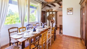Un coin repas avec une longue table en bois, dressée pour six personnes, entourée de chaises en bois. On y trouve une fenêtre avec des rideaux, un luminaire suspendu et une étagère fixée au mur, où sont rangés des ustensiles de cuisine.