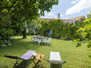 Un jardin avec une table dressée pour le repas, entouré de verdure et bordé d’un bâtiment dont le toit est recouvert de tuiles.