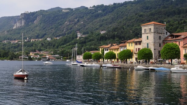 Une ville située au bord de l’eau, où des bateaux sont amarrés le long de la rive, avec un imposant immeuble en forme de tour et, en arrière-plan, des collines verdoyantes.
