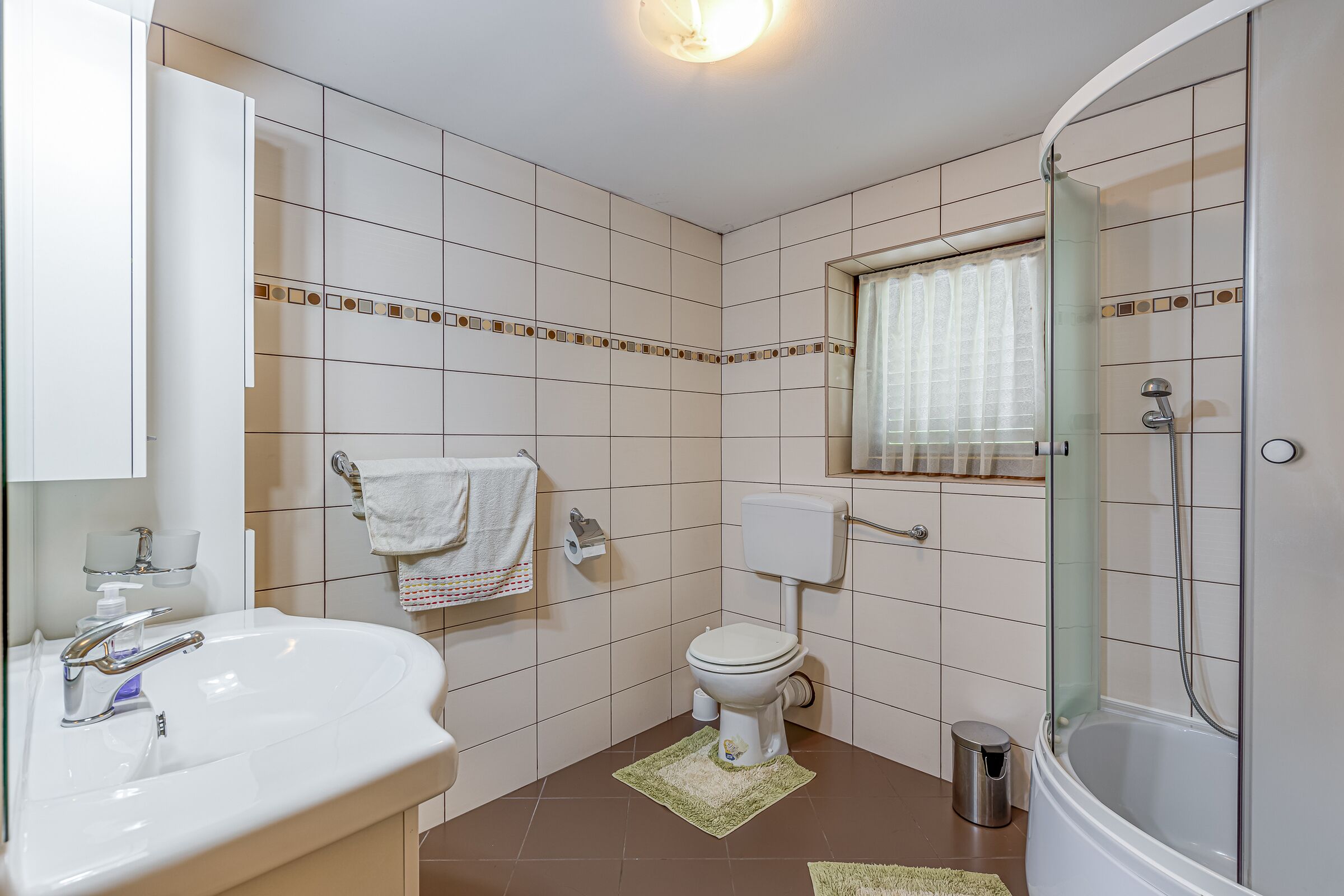 A bathroom with a sink, toilet, shower, and a window with a curtain.