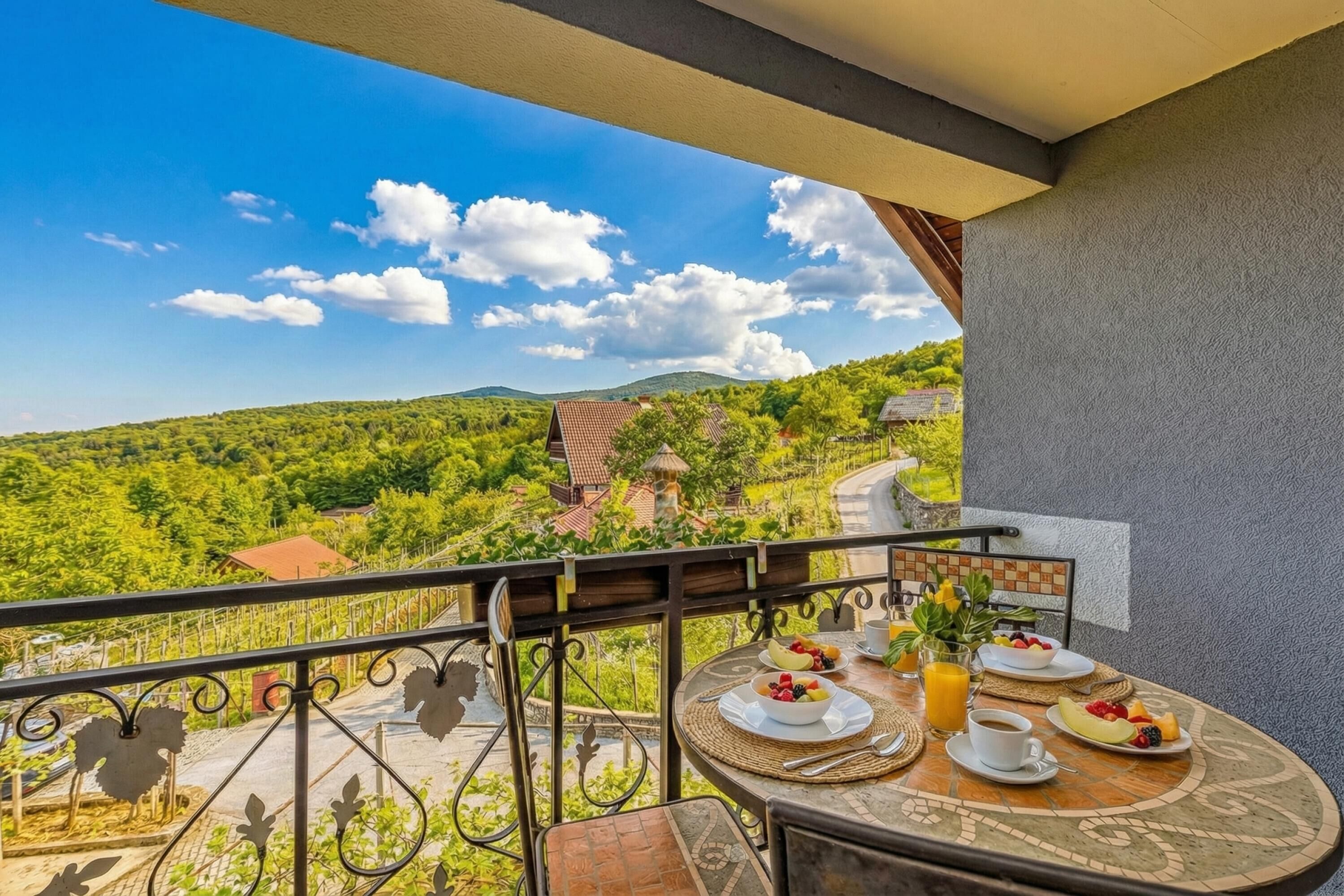 Un balcón con una mesa dispuesta para el desayuno, con vistas a un paisaje exuberante con árboles y un edificio distante.