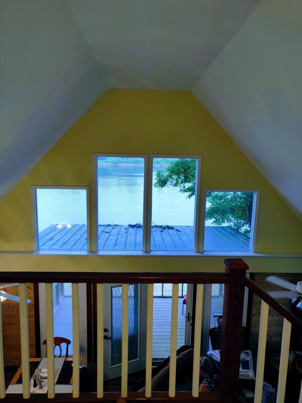 Ein Zimmer mit schrägem Dach und drei Fenstern mit Blick auf eine Holzterrasse und Wasser.