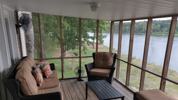 A covered porch with a wicker sofa, a chair, and a table, overlooking a lake. - Riverfront Retreat & Fisherman's Gold, on TN River (Savannah)
