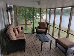 Ein überdachter Balkon mit einem Korbsessel, einem Sessel und einem Tisch, mit Blick auf einen See.