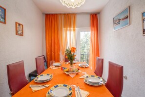 A dining room with a table set for four, orange curtains, and a view of greenery outside.